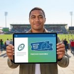 Un homme souriant présente une tablette affichant un billet de match de rugby officiel dans un stade en arrière-plan.