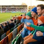 Une famille souriante composée de deux parents et deux jeunes enfants avec des bonnets regarde un match de rugby.