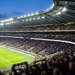 Vue panoramique d'un stade de football rempli de supporters lors d'un match en soirée sous les projecteurs.