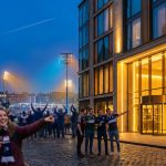 Des supporters de rugby joyeux devant un hôtel moderne à Édimbourg, avec un stade illuminé en arrière-plan au crépuscule.